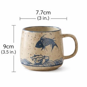 May include: A beige ceramic mug with a blue fish and wave design. The mug is 3.5 inches tall and 3 inches wide. It has a rounded shape and a large handle. The mug has a speckled finish.
