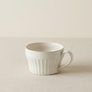 Peut inclure: Une tasse en céramique blanche avec une anse incurvée. La tasse présente des détails verticaux côtelés sur la moitié inférieure. La tasse est posée sur une surface de couleur neutre.