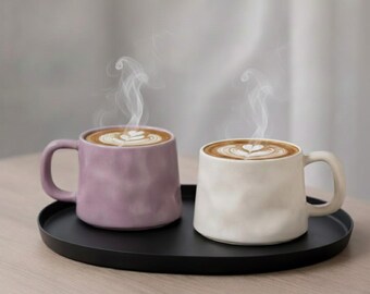 Peut inclure: Deux tasses en céramique, une lavande et une crème, remplies de café et de latte art, posées sur un plateau noir. De la vapeur s'élève du café. Les tasses ont une surface texturée et un design simple.