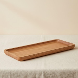 Può includere: Un vassoio rettangolare in legno con finitura marrone chiaro. Il vassoio ha un'area incassata poco profonda e bordi arrotondati. È posizionato su una superficie bianca e strutturata.