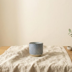 Peut inclure: Une tasse en céramique avec un motif à rayures bleues et blanches. La tasse est cylindrique avec une base beige et repose sur un tissu en lin beige. L'arrière-plan est de couleur beige clair.