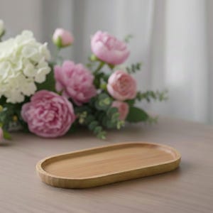 Può includere: Un vassoio ovale in legno marrone chiaro. Il vassoio ha una superficie liscia e un bordo leggermente rialzato. Sullo sfondo ci sono fiori rosa e bianchi.