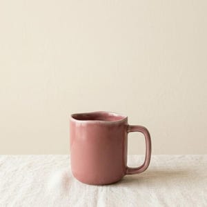 Könnte beinhalten: Eine staubrosa Keramiktasse mit rundem Korpus und einfachem Henkel. Die Tasse hat einen leicht unregelmäßigen Rand und steht auf einer weißen Leinenoberfläche. Der Hintergrund ist einfarbig und neutral.
