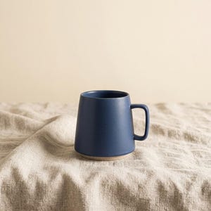 May include: A matte navy blue ceramic mug with a tapered silhouette and a single handle. The base of the mug features a contrasting light beige unglazed rim, providing a distinct visual separation from the smooth, dark blue body of the vessel.