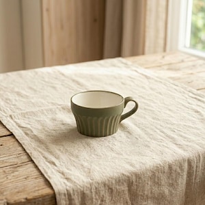 Könnte beinhalten: Eine grüne Keramik-Teetasse mit gerippter Außenseite und cremefarbenem Innenraum steht auf einer beigefarbenen Leinentischdecke. Die Tasse hat einen gebogenen Henkel und steht auf einem rustikalen Holztisch, was eine gemütliche Atmosphäre suggeriert.