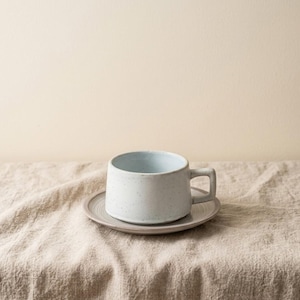 Puede incluir: Un juego de taza y platillo de cerámica. La taza es de color azul claro con una textura moteada y un asa. El platillo es de un color a juego. El juego está colocado sobre una superficie de lino beige.