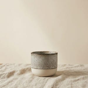 May include: A speckled gray ceramic mug with a cream-colored base. The mug has a cylindrical shape and a textured surface. The mug is sitting on a neutral-colored linen cloth.
