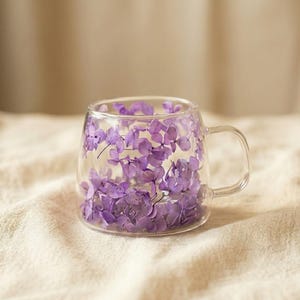 May include: A clear glass mug filled with delicate purple flowers. The mug has a handle and is set against a neutral-toned fabric background, creating a soft and inviting aesthetic. The flowers add a touch of natural beauty.