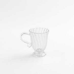 May include: A white ceramic mug with a black handle and the text "But First, Coffee" in a black, handwritten-style font. The mug is on a white surface.