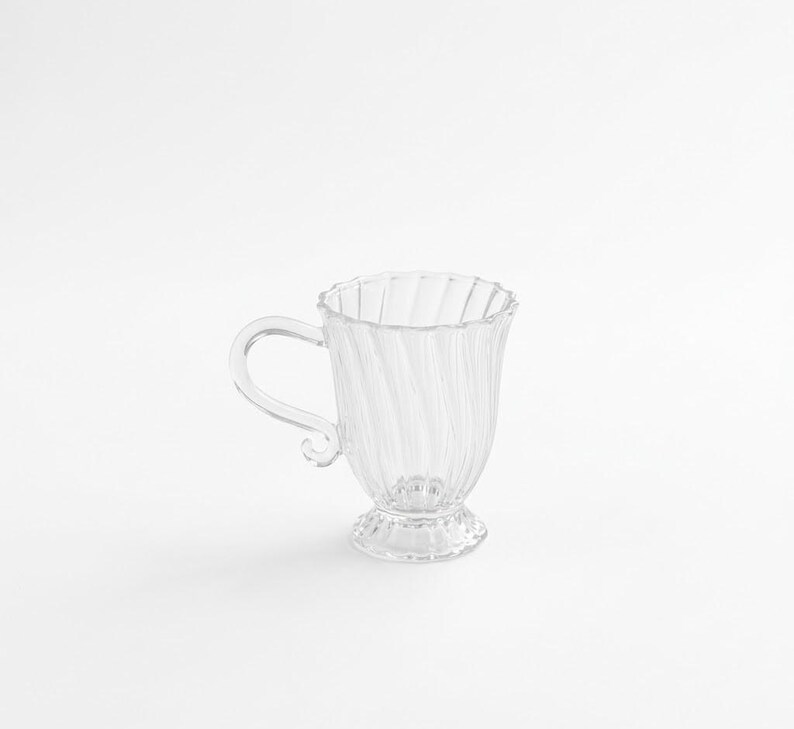 May include: A white ceramic mug with a black handle and the text "But First, Coffee" in a black, handwritten-style font. The mug is on a white surface.