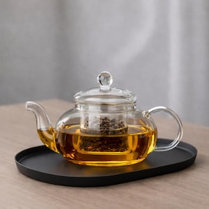 May include: A clear glass teapot with a glass infuser filled with tea leaves. The teapot is filled with amber-colored tea and has a curved handle and spout. It sits on a black oval tray.