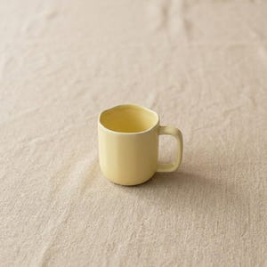 May include: A pale yellow ceramic mug with a rounded body and a handle. The rim of the mug has a slightly wavy design. The mug is empty and sits on a beige, textured surface.