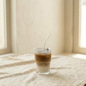 May include: A clear glass with a lid and a curved glass straw, filled with a layered drink of light and dark brown liquids. The glass sits on a textured, off-white surface near a window.