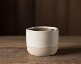 May include: A small, two-toned ceramic coffee cup filled with dark liquid, likely coffee. The upper portion is a creamy white, while the bottom is a light beige. The cup sits on a wooden surface, with a blurred brown background.