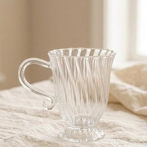 May include: A clear glass mug with a curved handle and a ridged design. The mug has a flared rim and sits on a small, ornate base. The transparent glass allows light to pass through. Suitable for serving hot drinks, it is a decorative item.