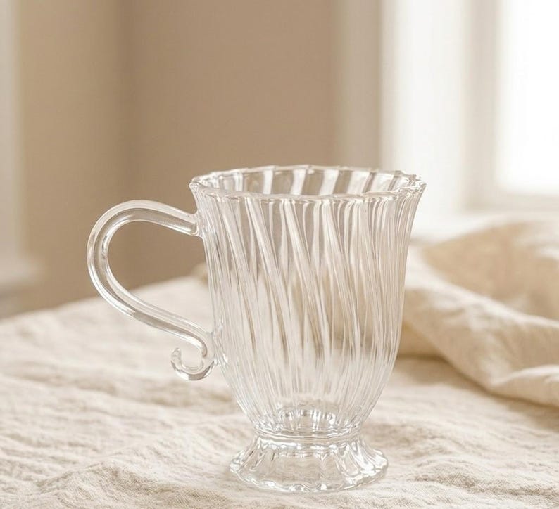 May include: A clear glass mug with a curved handle and a ridged design. The mug has a flared rim and sits on a small, ornate base. The transparent glass allows light to pass through. Suitable for serving hot drinks, it is a decorative item.