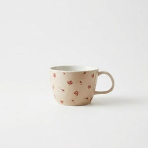 May include: A beige ceramic mug with a white interior and a handle. The mug is adorned with a pattern of small, red floral designs. The mug is a standard size for coffee or tea.
