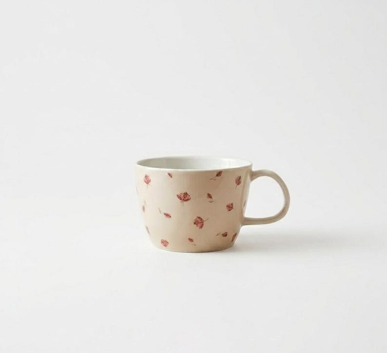 May include: A beige ceramic mug with a white interior and a handle. The mug is adorned with a pattern of small, red floral designs. The mug is a standard size for coffee or tea.
