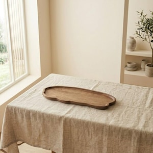 Puede incluir: Una bandeja de madera para servir con una forma orgánica única descansa sobre un mantel de lino beige. La bandeja es de color marrón cálido y parece estar hecha de madera maciza. La habitación tiene una paleta de colores neutros con una ventana y una estantería decorativa en el fondo.