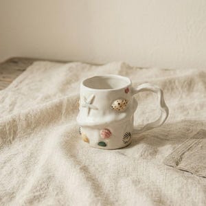 Könnte beinhalten: Weißer Keramikbecher, verziert mit Muscheln und einem Seestern. Der Becher hat einen wellenförmigen Griff und ist mit verschiedenen Muscheldesigns in Rosa-, Gold- und Grüntönen verziert. Der Becher steht auf einem beigen Leinentuch.