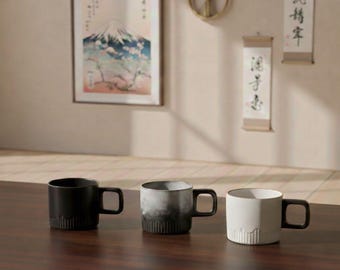 May include: Three ceramic mugs in black, gray, and white with black handles sit on a dark wooden surface. The mugs have a minimalist design with vertical ridges at the base. The background features Japanese-style art and decor.