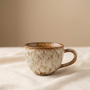 Pode incluir: Uma chávena de café de cerâmica com um exterior castanho claro texturizado e uma borda e pega castanhas mais escuras. A chávena assenta sobre um tecido creme, com um fundo neutro. A chávena tem um design único e orgânico.