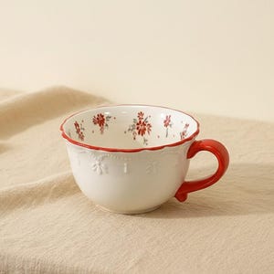 May include: A white ceramic teacup with a red handle and rim. The cup features a floral pattern in red and white, with embossed details. The cup is set against a neutral background.