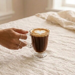 May include: A clear glass coffee mug filled with dark coffee and topped with a heart-shaped design in the foam. The mug has a decorative handle and sits on a pedestal base. The image is set on a neutral-coloured, textured surface.