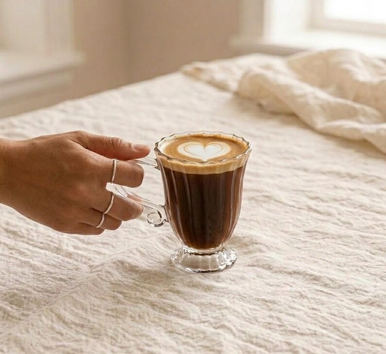 May include: A clear glass coffee mug filled with dark coffee and topped with a heart-shaped design in the foam. The mug has a decorative handle and sits on a pedestal base. The image is set on a neutral-coloured, textured surface.