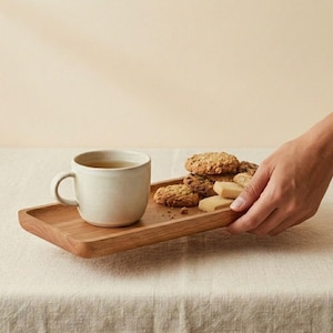 Pu&ograve; includere: Un vassoio di legno con una tazza di ceramica bianca piena di t&egrave; e un assortimento di biscotti. Una mano raggiunge il vassoio, appoggiato su una tovaglia colorata neutra. I biscotti sono di varie forme.