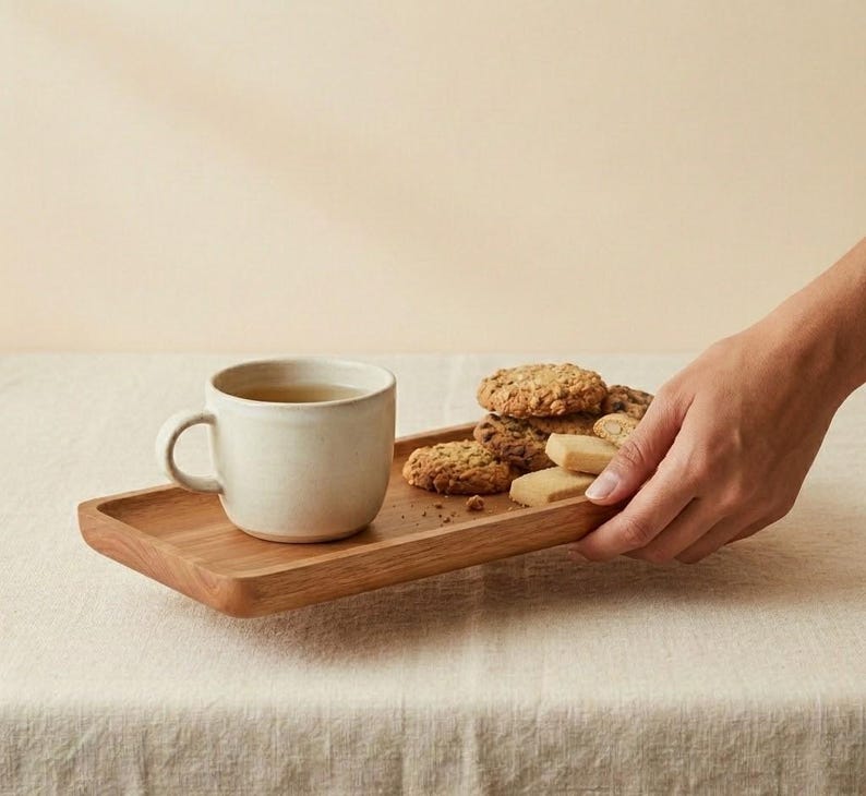 Pu&ograve; includere: Un vassoio di legno con una tazza di ceramica bianca piena di t&egrave; e un assortimento di biscotti. Una mano raggiunge il vassoio, appoggiato su una tovaglia colorata neutra. I biscotti sono di varie forme.
