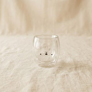 Könnte beinhalten: Ein klares, doppelwandiges Glas mit einem Bärengesicht-Design. Das Bärengesicht besteht aus schwarzen Punkten für die Augen und einer schwarzen Linie für Nase und Mund. Das Glas hat eine ovale Form.