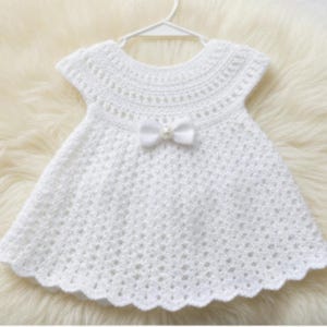 May include: A white, crocheted baby dress with a scalloped hem and short sleeves. A white bow with a pearl center adorns the front. The dress is displayed on a white hanger against a soft, white background.