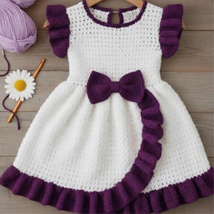 May include: A handmade white crochet dress with purple accents. The dress features a large bow, ruffled sleeves, and a ruffled hem. The dress is displayed on a wooden hanger.