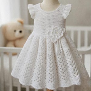 May include: A white crochet dress featuring a round neckline, short ruffled sleeves, and a flared skirt with a detailed openwork pattern. A large, dimensional crochet flower is attached to the waist, accented with a small dangling bead detail.