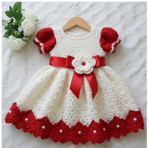 Puede incluir: Vestido de ganchillo hecho a mano en marfil y rojo, exhibido en una percha de madera. El vestido presenta una cinta roja, una flor blanca y bordes festoneados rojos. Las mangas son rojas con ribete blanco. Para bebé o niño pequeño.