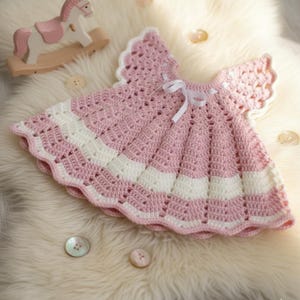 May include: A handmade, pink and white crochet baby dress with a scalloped hem and sleeves. The dress features a white ribbon bow at the neckline. The dress is lying on a white, fluffy surface.
