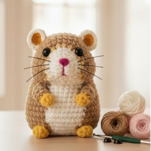Könnte beinhalten: Handgefertigtes gehäkeltes Hamsterspielzeug in Braun-, Weiß- und Gelbtönen. Der Amigurumi-Hamster hat schwarze Augen, Schnurrhaare und eine rosa Nase. Wollknäuel und ein Häkelhaken befinden sich im Hintergrund.
