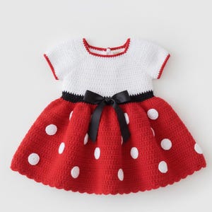 May include: A short-sleeved crochet dress featuring a white bodice and a red skirt with white polka dots. The waist is accented with a black satin ribbon bow. The neckline is trimmed in red, and the dress has a button closure at the back neck.