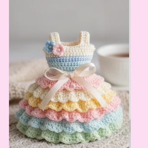 May include: A handmade crochet dress in pastel colors. The dress features a tiered skirt in shades of pink, yellow, and blue, with a cream-colored bodice and a delicate bow. A small flower embellishes the top. The dress is a decorative item.