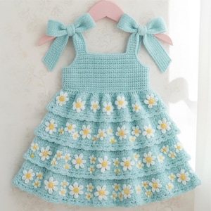 May include: A light blue crochet dress featuring three tiered ruffles and white daisy appliqués with yellow centers. The bodice has a square neckline and two shoulder straps that tie into bows at the top, hanging on a pink plastic hanger.