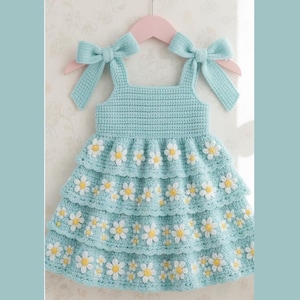 May include: A light blue crochet dress featuring three tiered ruffles adorned with white and yellow daisy flower appliques. The bodice has a square neckline and two wide shoulder straps that tie into bows at the top of the garment.