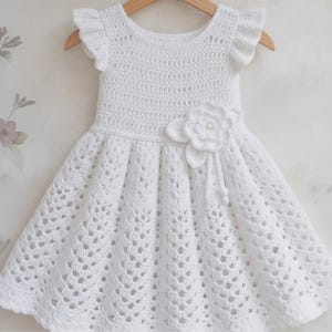 May include: A white crochet dress featuring a round neckline, short ruffled sleeves, and a flared skirt with a detailed openwork pattern. A large, layered crochet flower with a central bead accent is attached to the waistline of the garment.