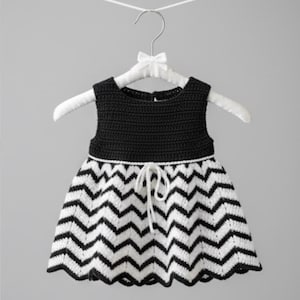 Puede incluir: Vestido de bebé de ganchillo en blanco y negro, colgado de una percha blanca. La parte superior es negra lisa y la falda presenta un estampado de chevrón. Un lazo blanco ciñe la cintura. El vestido es sin mangas.