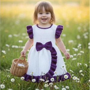 May include: A white crochet dress with purple accents, including a large bow and ruffled trim. The dress features a round neckline and short sleeves with ruffled edges. A small woven basket is held in the hand.
