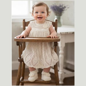 May include: A smiling baby wearing a cream-colored, sleeveless crochet dress and matching booties sits in a wooden high chair. The dress features a floral embellishment and a scalloped hem. The baby is looking at the camera.