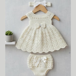 May include: A cream crochet baby outfit comprising a dress, bloomers, and a headband. The dress has a bow and a scalloped hem. The set is displayed on a wooden hanger. The bloomers also feature a bow. The background is neutral.