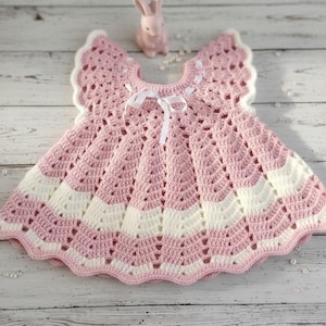May include: A handmade crochet baby dress in pink and white. The dress has a scalloped hem and sleeves, with a white ribbon bow at the neckline. A small pink bunny figurine is in the background.