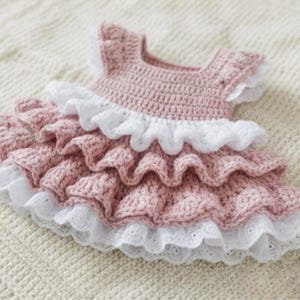 May include: A handmade, pink crochet dress with white ruffled trim. The dress features a round neckline and short sleeves. The skirt has multiple layers of ruffles, creating a textured and delicate appearance. The dress is designed for a baby or toddler.