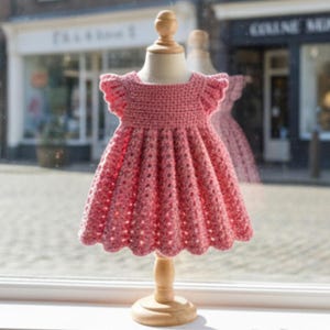 Puede incluir: Vestido rosa de ganchillo hecho a mano para niña, expuesto en un maniquí de madera. El vestido presenta un dobladillo festoneado, mangas con volantes y un corpiño texturizado. El vestido se exhibe frente a una ventana.